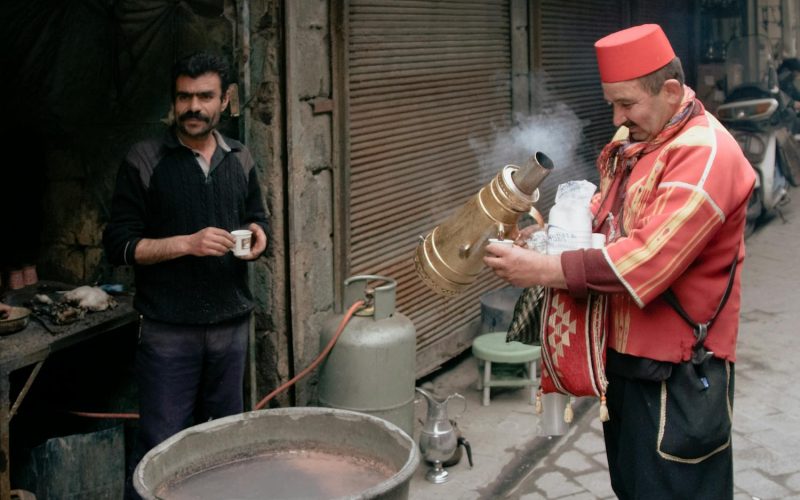 A vendor in traditional clothing serves a drink in an urban alley setting.