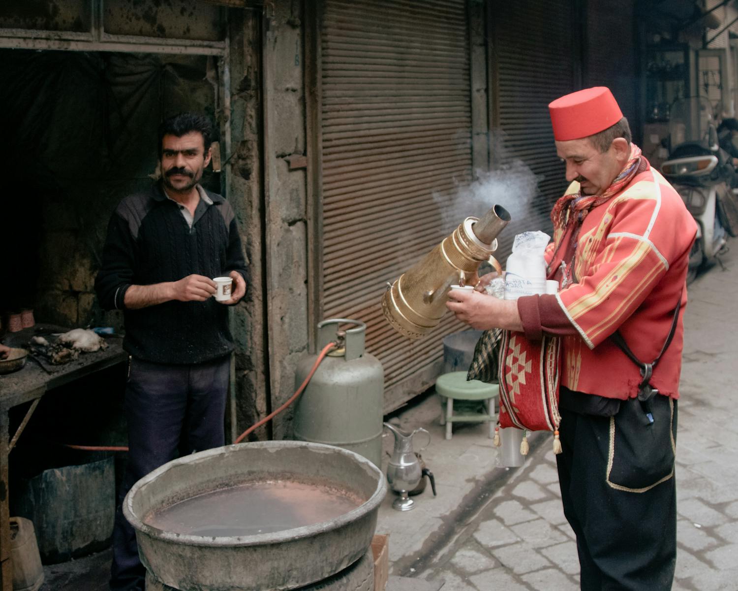 A vendor in traditional clothing serves a drink in an urban alley setting.